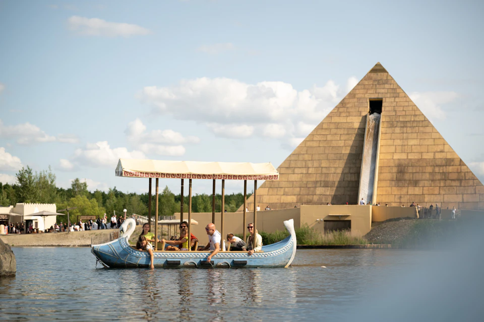 A group of people ride a decorated boat on a waterway near a large pyramid structure with a waterfall feature.