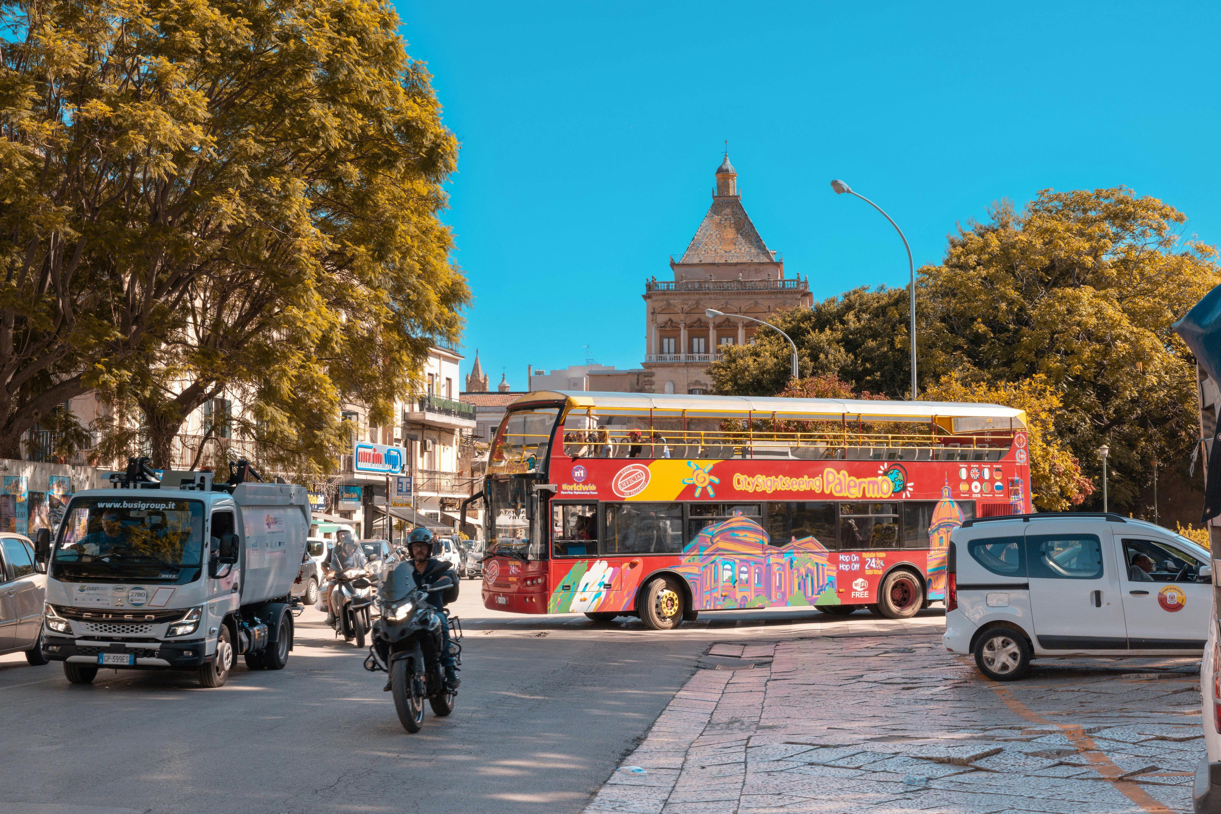 Un colorato autobus turistico a due piani in una strada urbana trafficata con motociclette, alberi e un edificio storico sotto un cielo limpido.