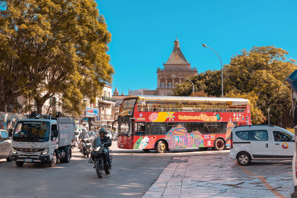 A colorful double-decker tourist bus in a busy urban street with motorcycles, trees, and a historic building under a clear sky.