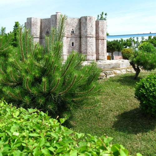 Un castello in miniatura circondato da una vegetazione lussureggiante, cespugli e piccoli pini sotto un cielo azzurro e limpido.