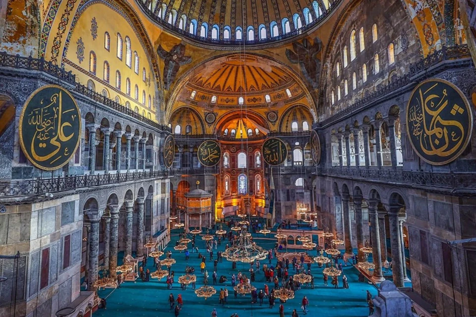 Interior of a large historic building with domed ceiling, ornate decorations, and people walking on a blue floor.
