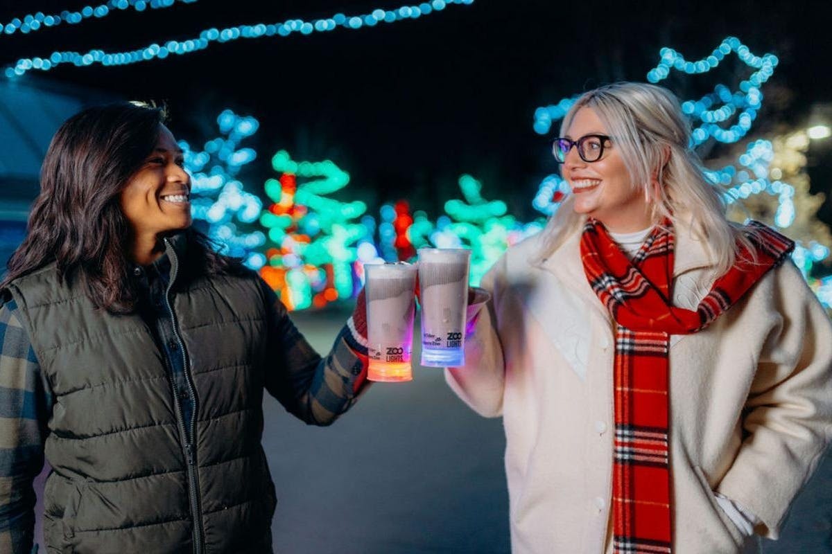 흰색 코트와 빨간색 스카프를 두른 여성이 축제 분위기의 밝은 야외 공간에서 음료를 들고 미소를 짓고 있습니다.