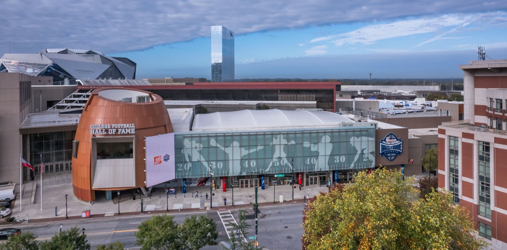 Budova College Football Hall of Fame v Atlantě s nedalekou moderní architekturou a výhledem na ulici se stromy a několika chodci.
