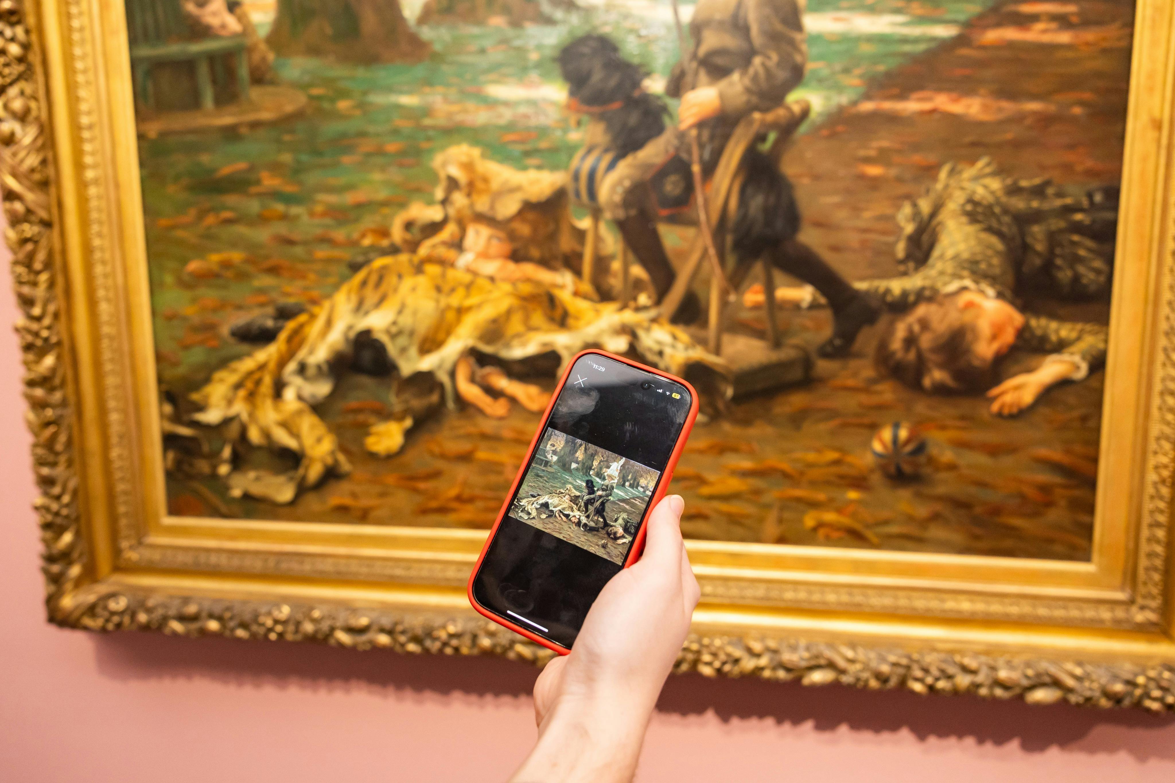 미술관에 전시된 그림을 배경으로 그림의 이미지가 담긴 스마트폰을 들고 있는 사람.