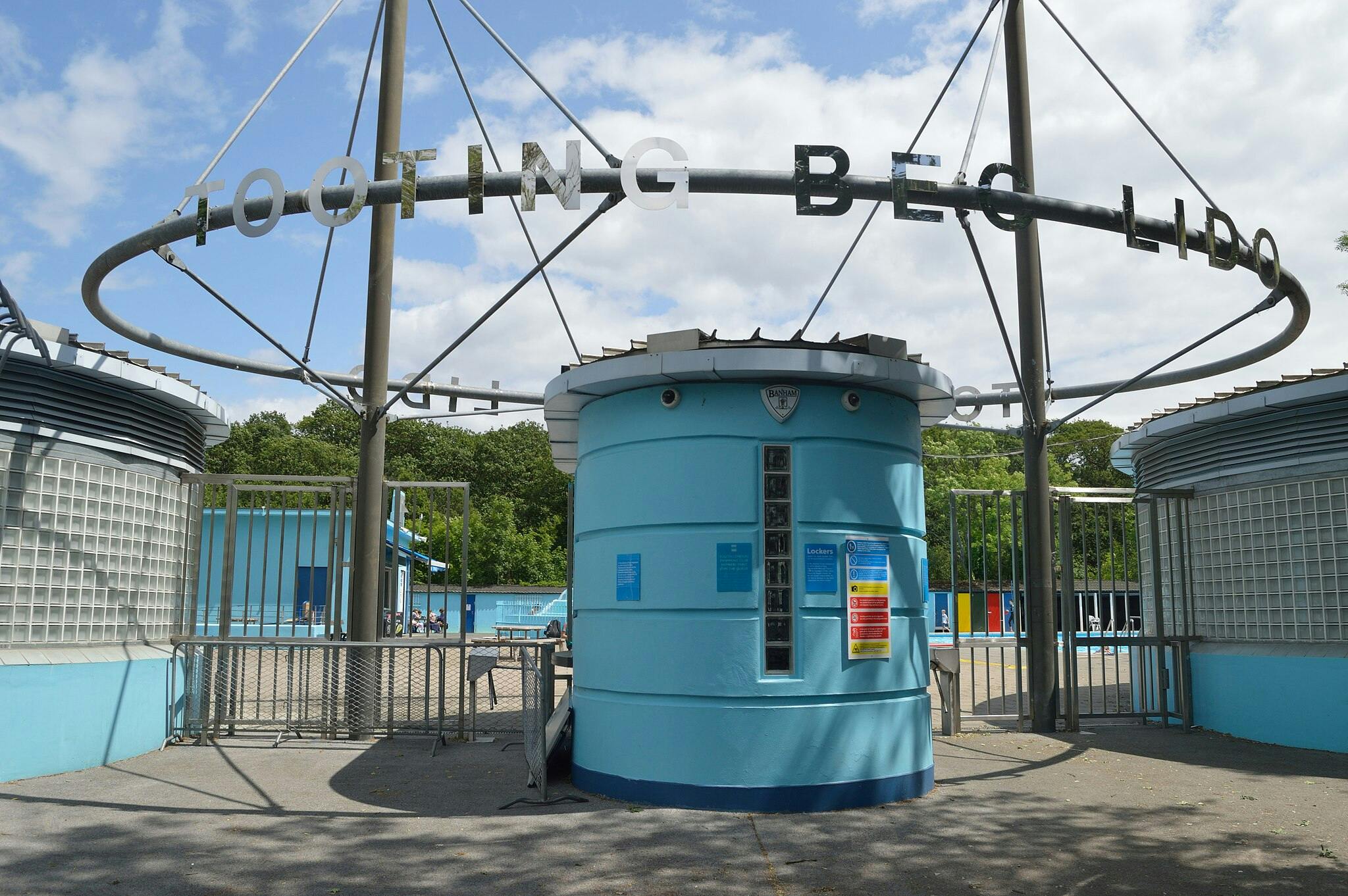 Eingang zum Strandbad Tooting Bec mit einem blauen, runden Gebäude mit Informationsschildern, einem Eingangstor und einem Schild mit der Aufschrift "TOOTING BEC LIDO".