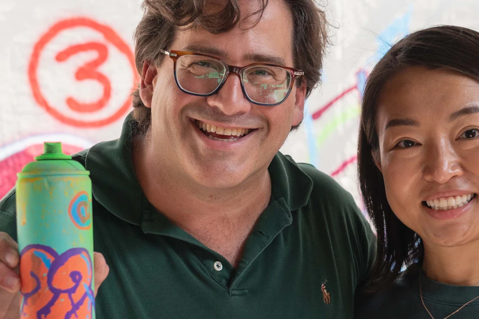 Two people smiling, holding colorful spray paint cans, standing in front of a wall with vibrant graffiti art.