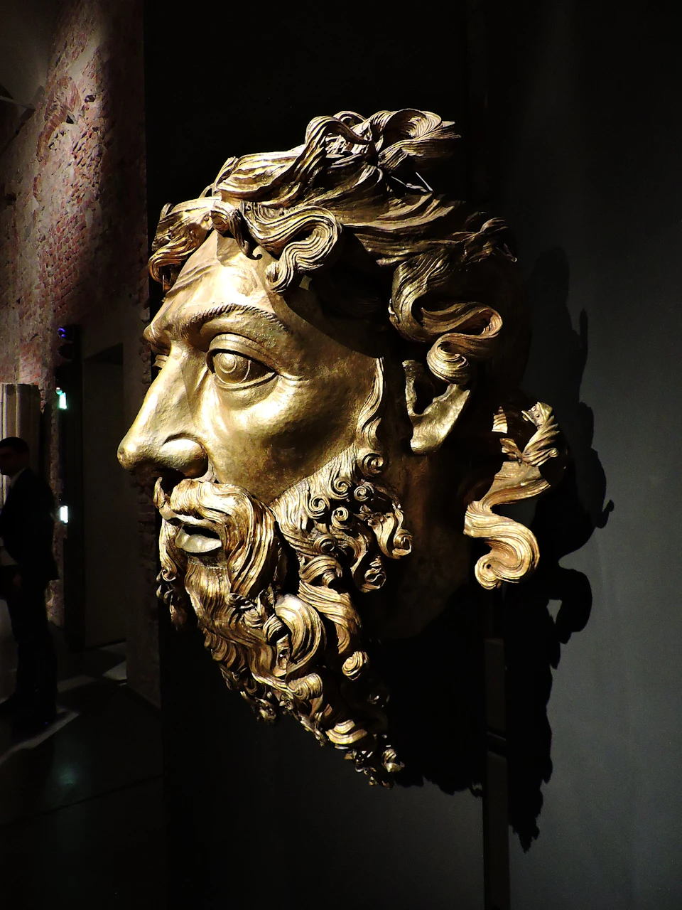 A large, intricately carved golden face with curly hair and a beard displayed on a wall in a dimly lit room.