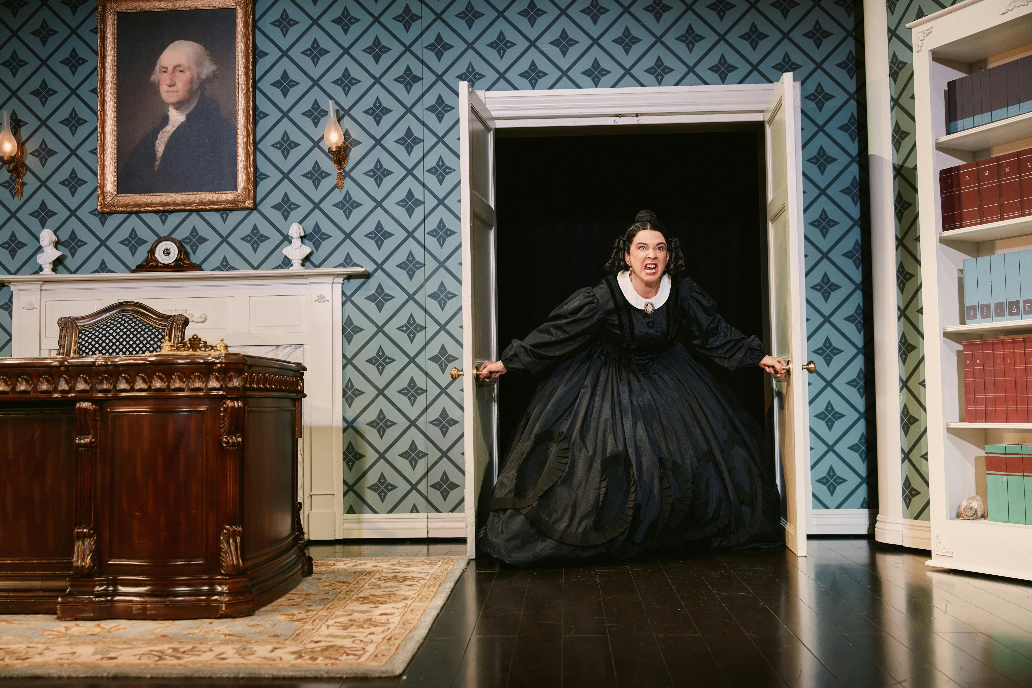 A woman in a black dress stands dramatically in a doorway of an ornate room with a portrait of George Washington on the wall.