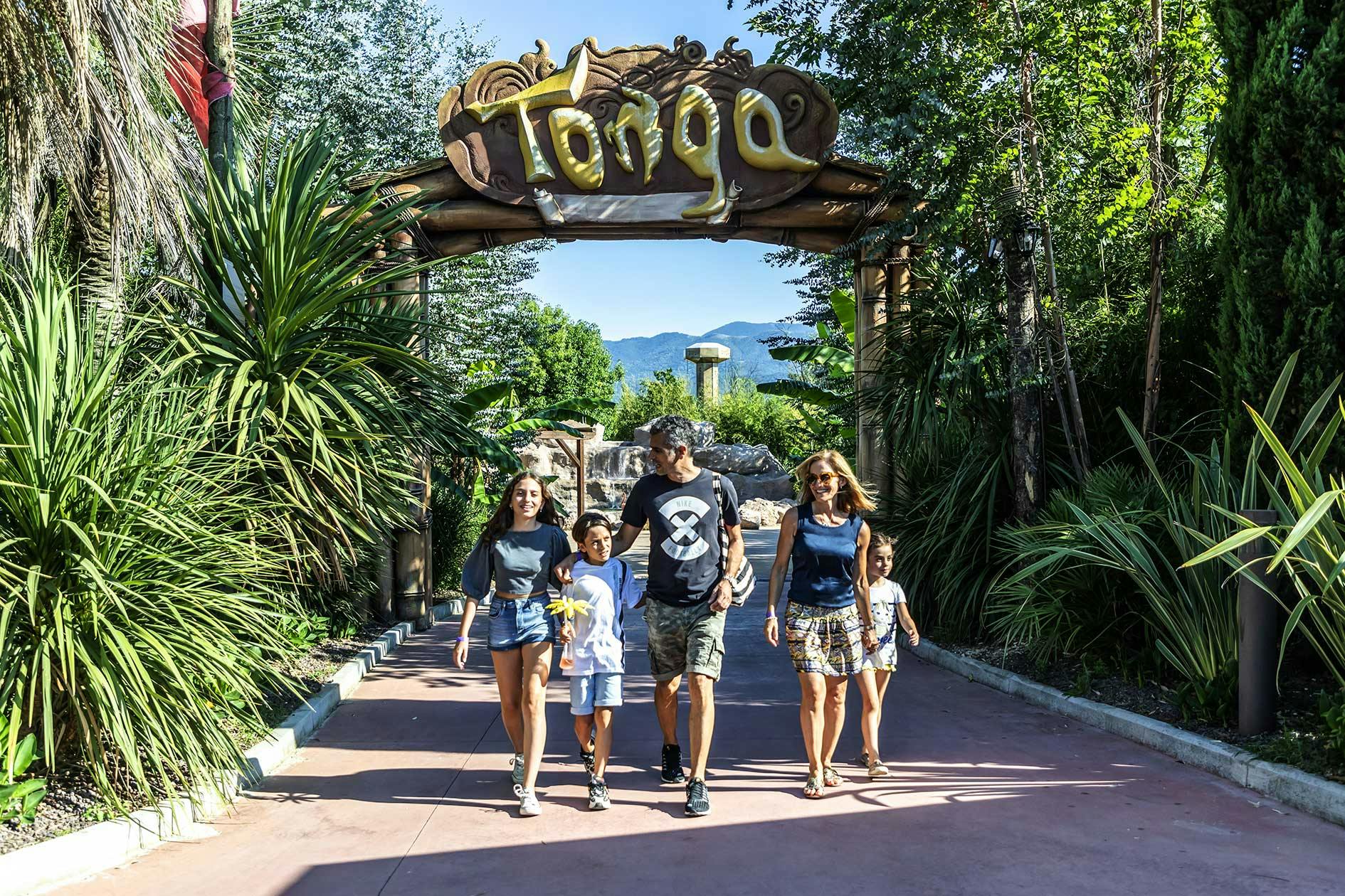 Μια πενταμελής οικογένεια περπατά κάτω από μια μεγάλη ξύλινη αψίδα με την ένδειξη "Tonga" σε ένα καταπράσινο τροπικό περιβάλλον.