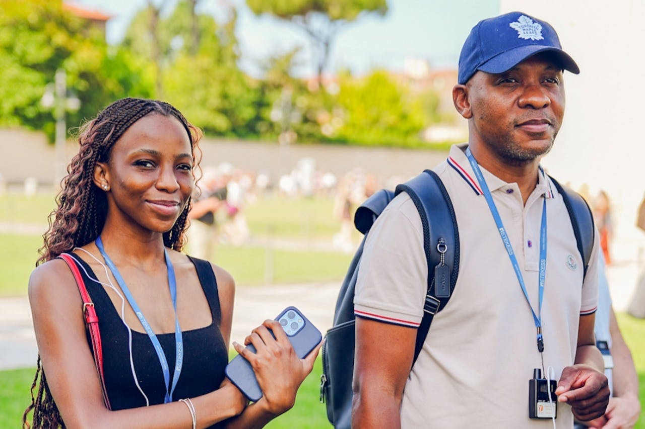 Two individuals with lanyards and badges, one holding a phone, outdoors with greenery and blurred people in the background.
