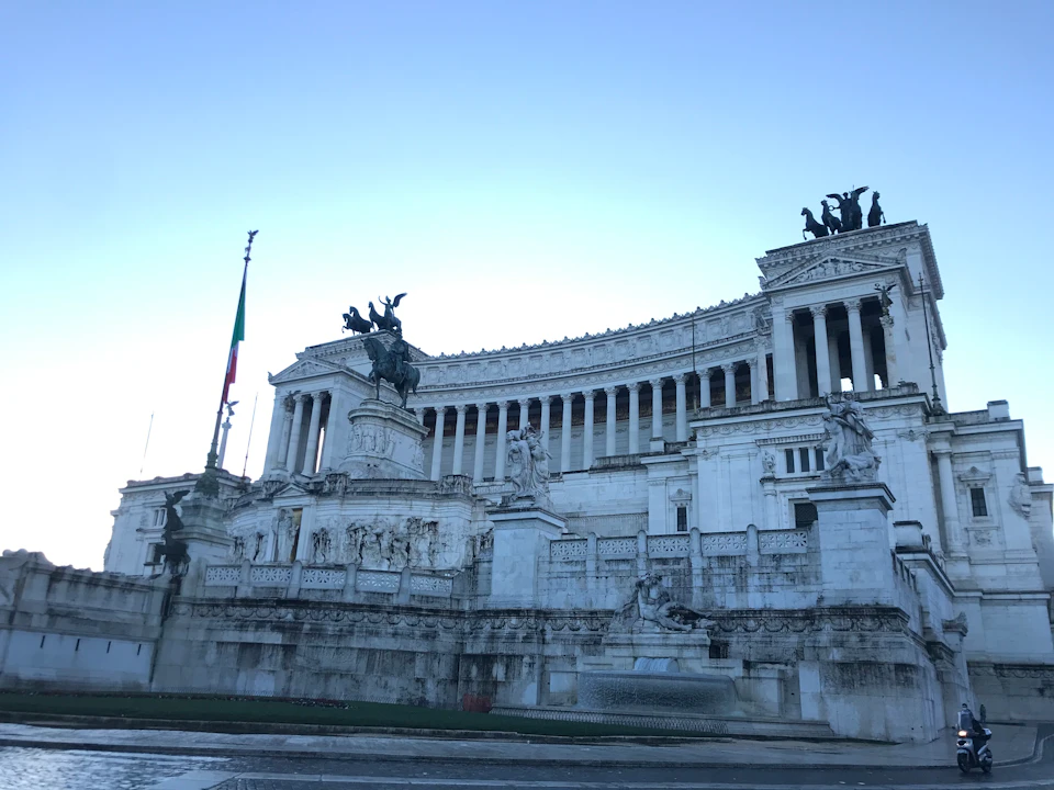White neoclassical building with statues, columns, and a mounted horseman. Italian flag on the left and clear sky above.