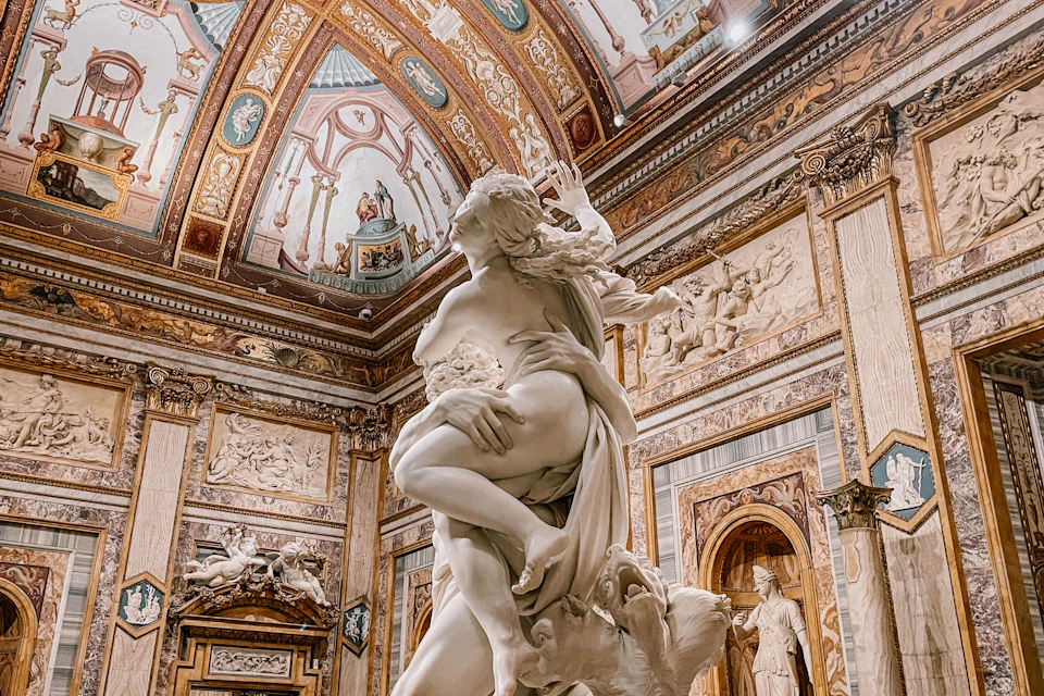 A marble sculpture depicting an embracing couple in a lavishly decorated museum hall with ornate ceilings and walls.