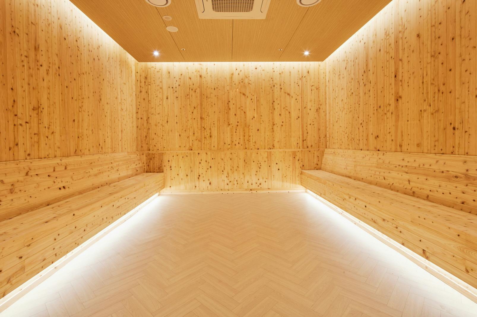A spacious room with light wooden walls and benches, illuminated by recessed lighting, with a light herringbone-patterned floor.