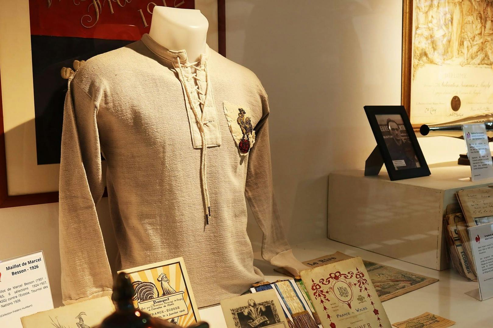Museum display featuring a beige laced shirt with an emblem, vintage documents, and a framed photo in the background.