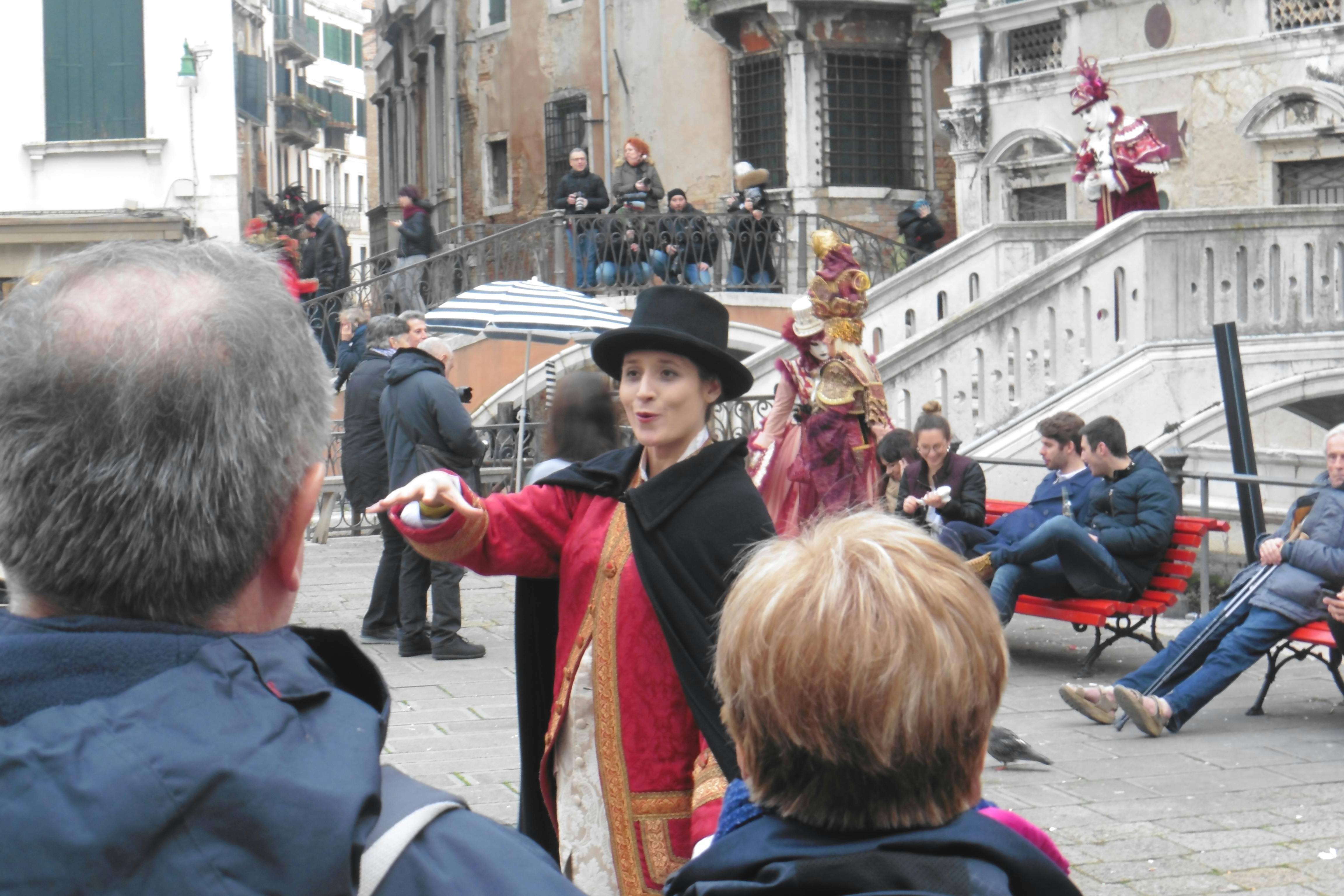 歴史的なコスチュームとシルクハットを着たパフォーマーが、階段と人々を背景にした装飾されたヨーロッパの広場で、少人数の観客を魅了しています。