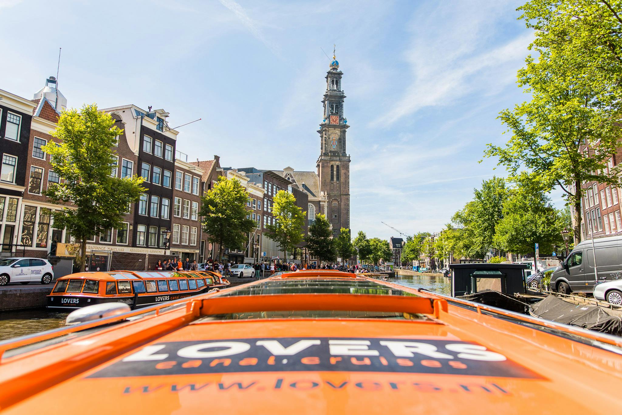 Orangefarbenes Tour-Boot auf einer Amsterdamer Gracht, mit hohen Gebäuden und einem Kirchturm im Hintergrund unter einem klaren blauen Himmel.