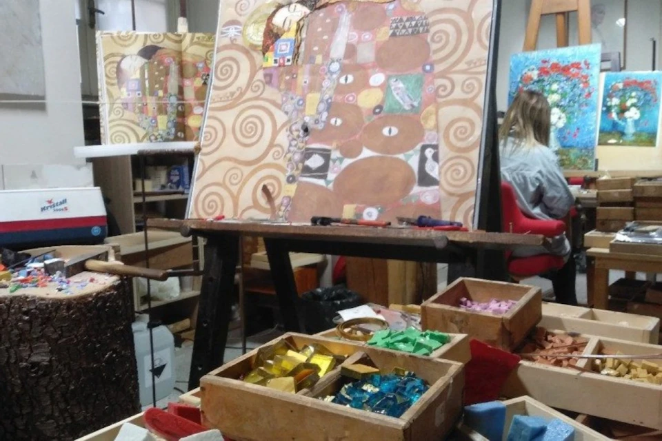 An art studio with a painting on an easel resembling Klimt's style and boxes of colorful material in the foreground.