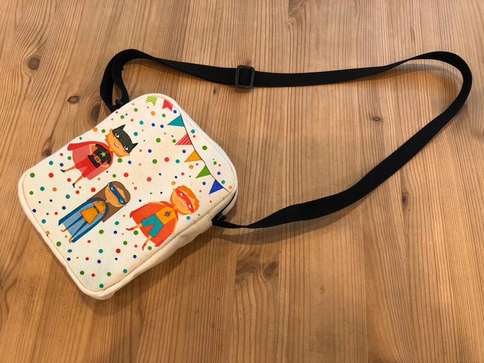 A small shoulder bag with a colorful design featuring cartoon superheroes and confetti, placed on a wooden surface.