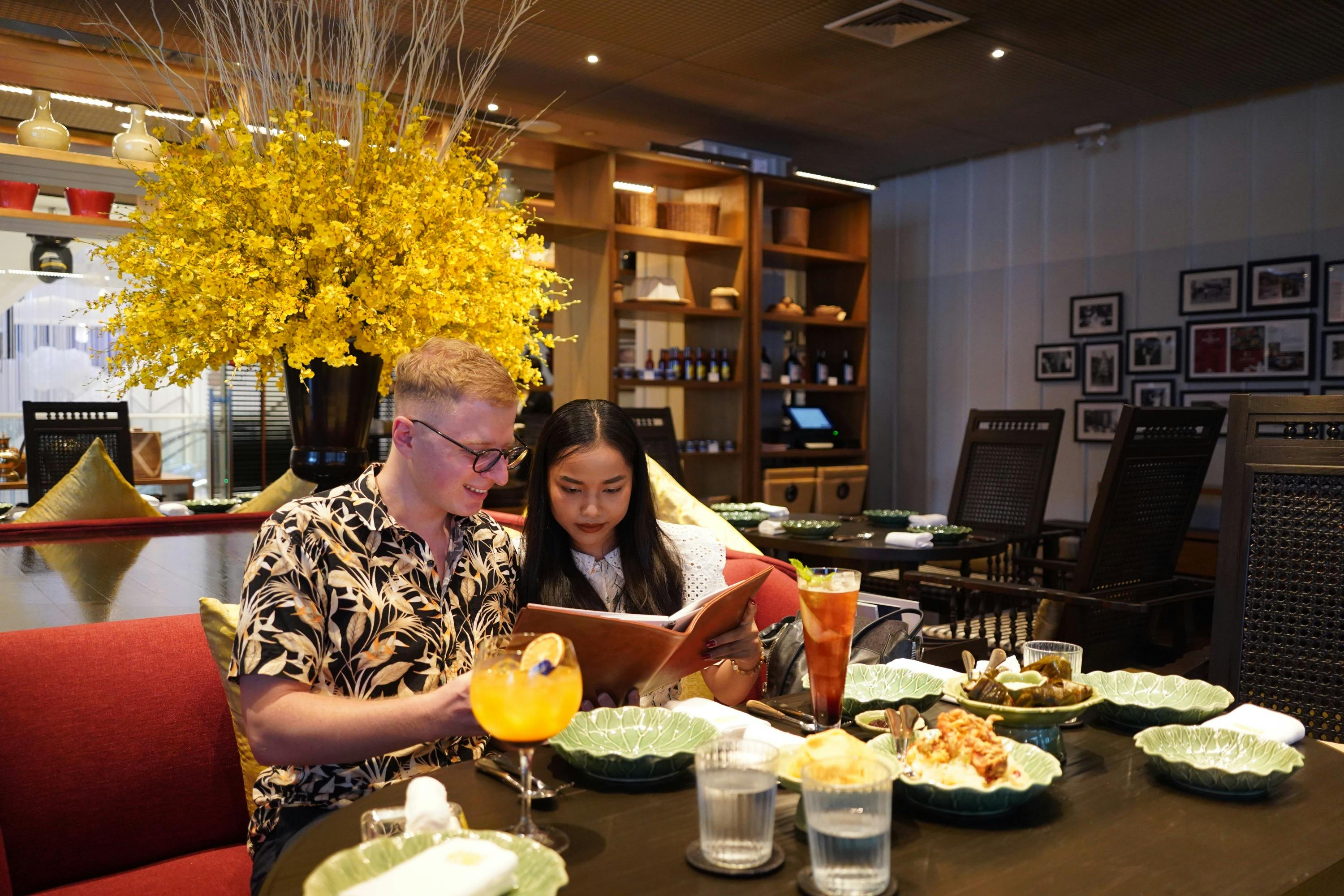 Two people sitting at a restaurant table with drinks and food, looking at a menu. A yellow floral arrangement is behind them.