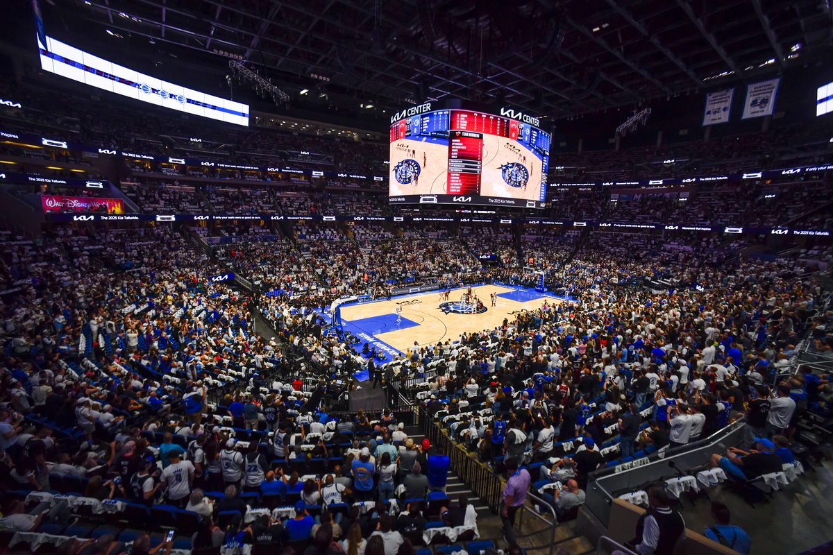 A crowded basketball arena during a game, with a large overhead scoreboard and fans filling the stands.