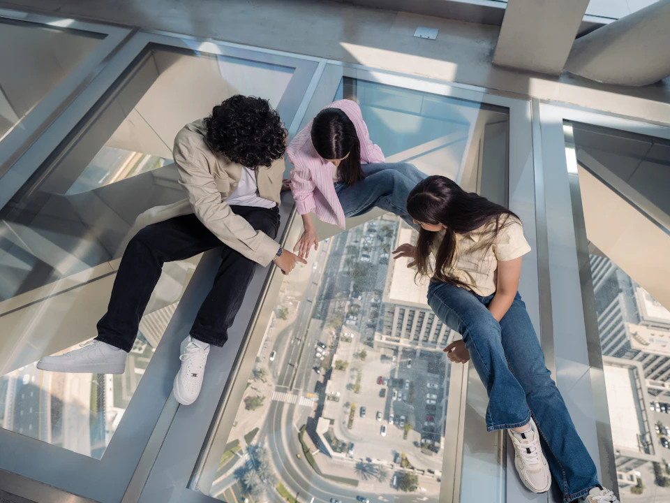 Three people sitting on a glass floor looking down at the city street and buildings below.