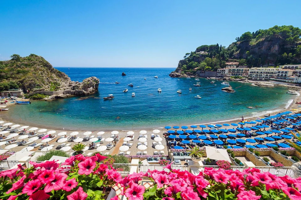 Coastal scene with clear blue sea, beach umbrellas, boats in the water, rocky cliffs, and vibrant pink flowers in the foreground.