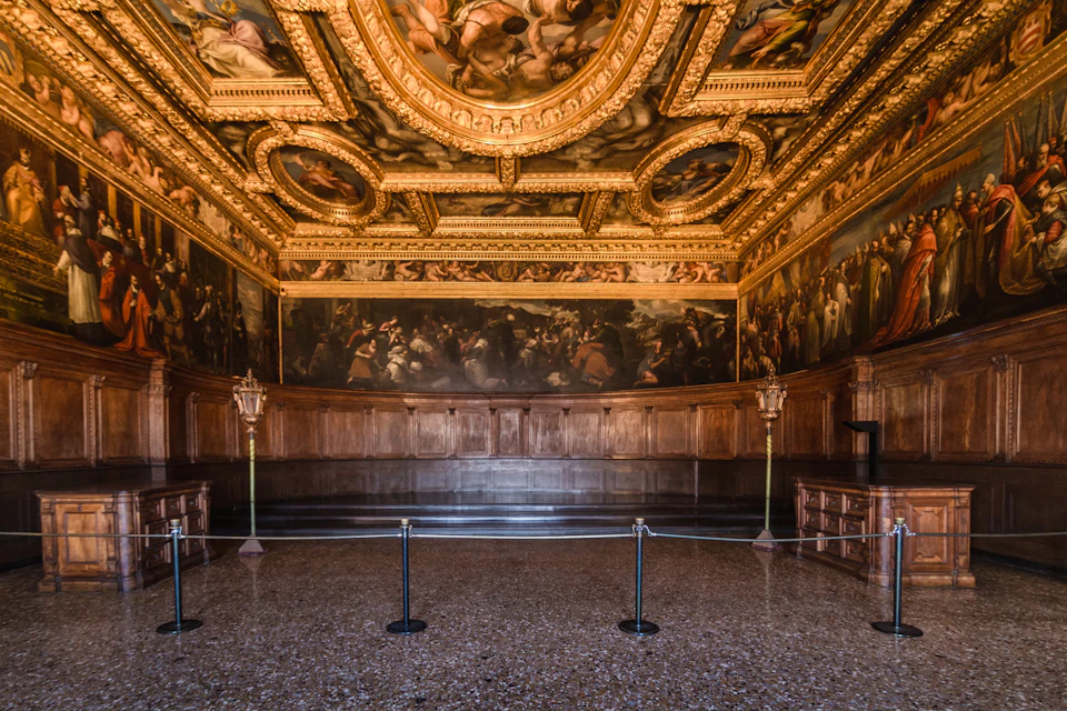 Lavish, ornate room with golden accents, detailed ceiling murals, dark wood paneling, and two barrier ropes in the foreground.