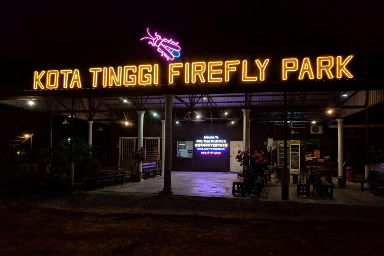 A night view of Kota Tinggi Firefly Park entrance with illuminated signage and seating areas under a roof.