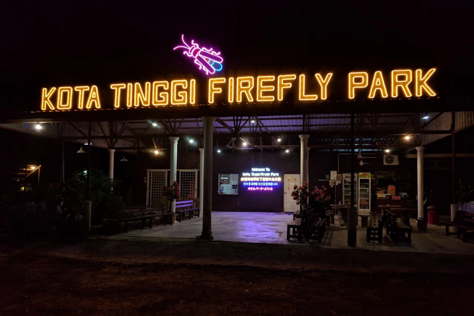 A night view of Kota Tinggi Firefly Park entrance with illuminated signage and seating areas under a roof.