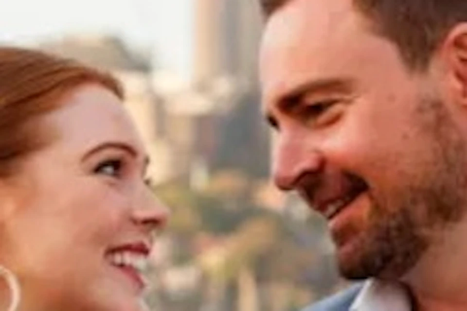 A smiling couple holding drinks on a boat with the Sydney Harbour Bridge in the background.