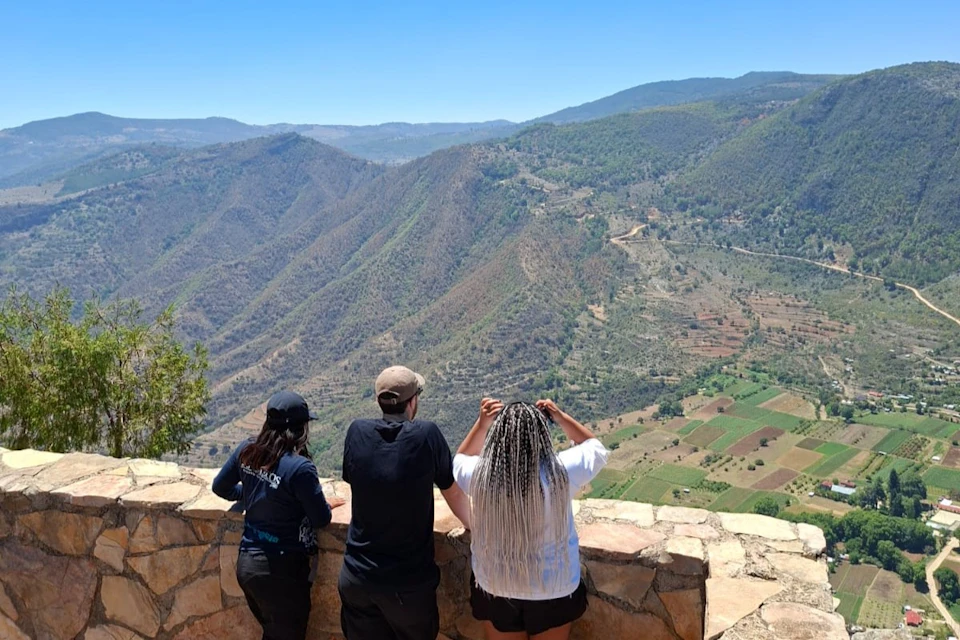 Three people standing by a stone railing, overlooking a scenic view of mountains and cultivated fields on a sunny day.