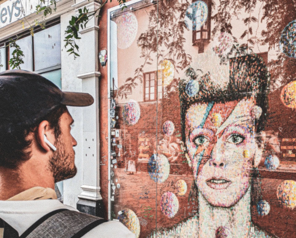 A man wearing a cap and earphones looks at a wall mural featuring a colorful portrait of a person with a lightning bolt on their face.