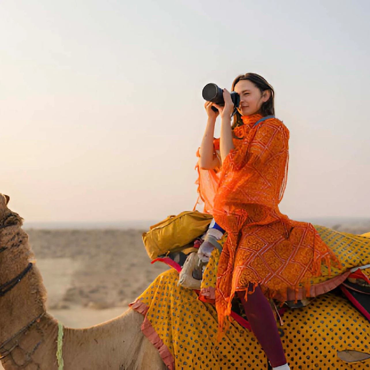 A woman in an orange shawl sits on a camel in a desert, using a camera with a long lens.