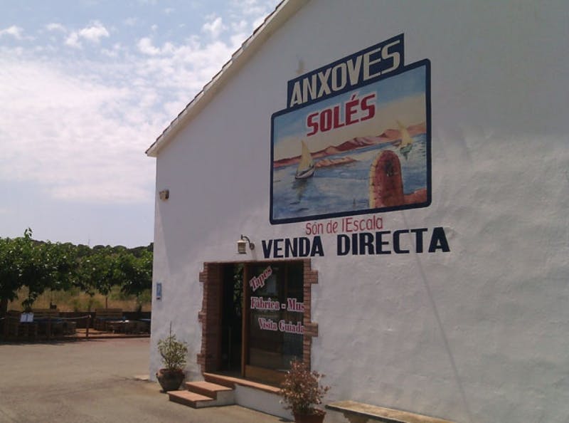 White building with large sign reading "ANXOVES SOLES" above a mural of boats. Entrance with potted plants and a tree on the left.