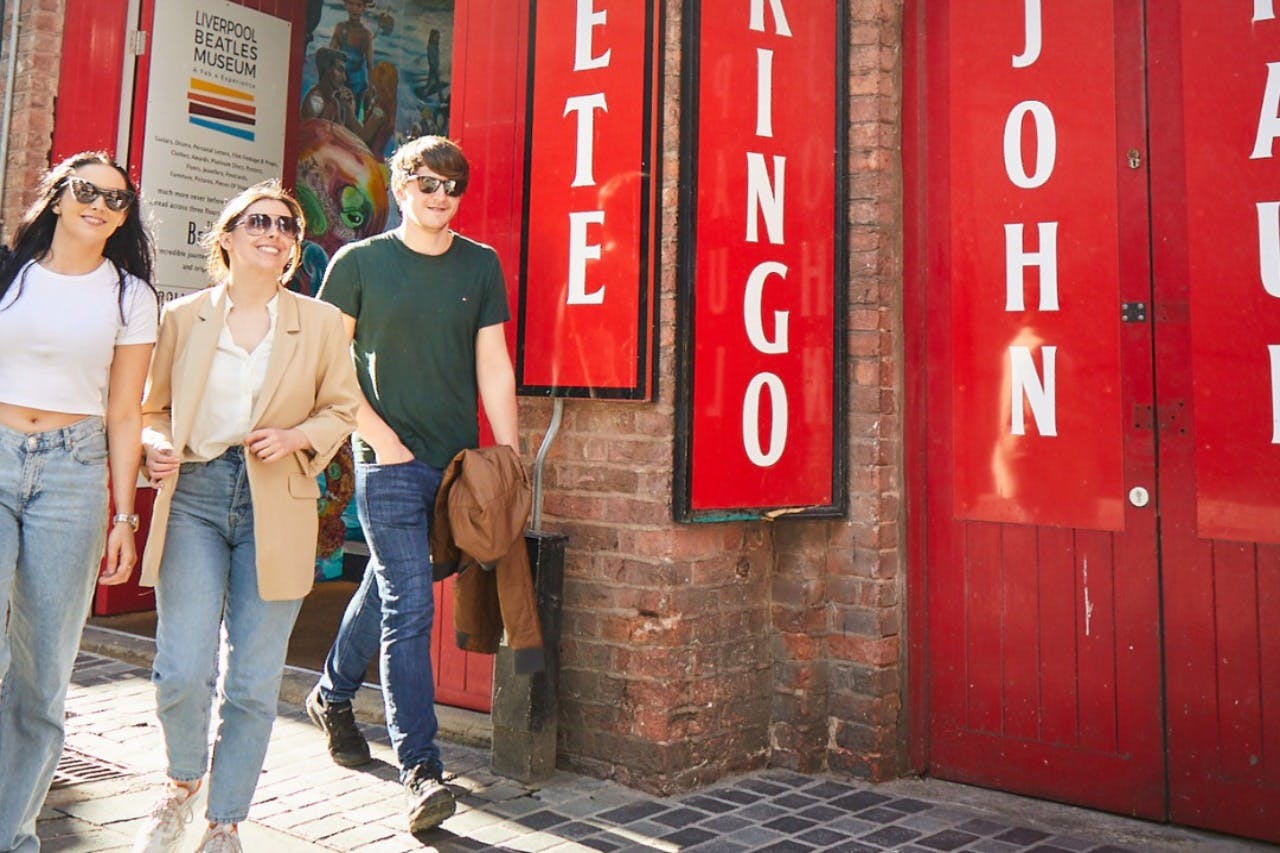 Tre persone che camminano davanti a un muro di mattoni con cartelli rossi con scritto "Pete" e "Ringo", vicino a un poster del Liverpool Beatles Museum.