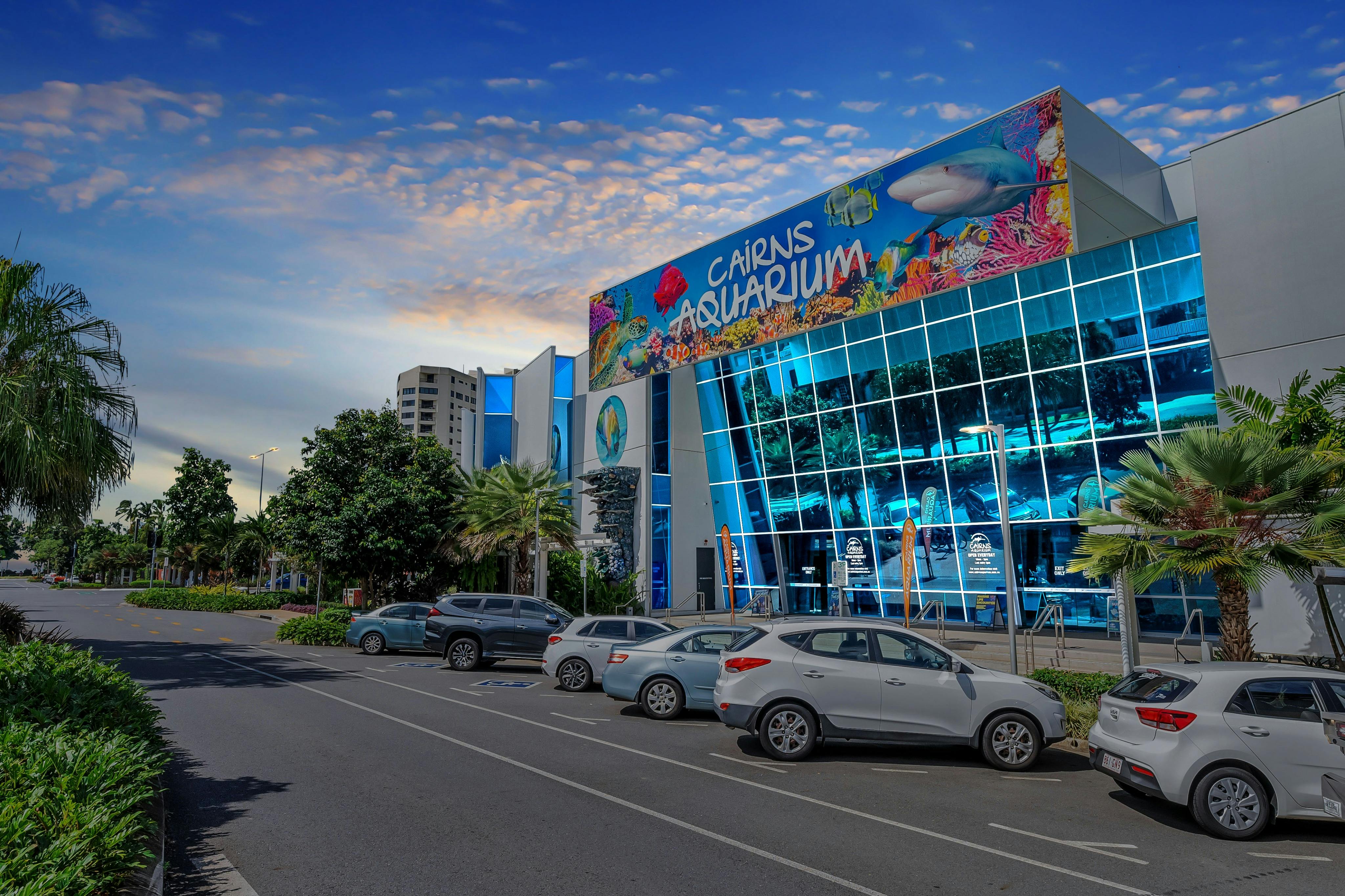 Ein modernes Gebäude mit der Aufschrift "Cairns Aquarium" mit großen Glasfenstern, parkenden Autos und Bäumen unter blauem Himmel.