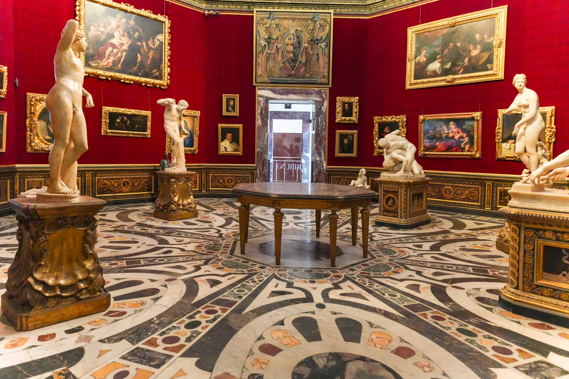 An ornate museum room with red walls, classical paintings, marble statues, and a central wooden table on a mosaic floor.