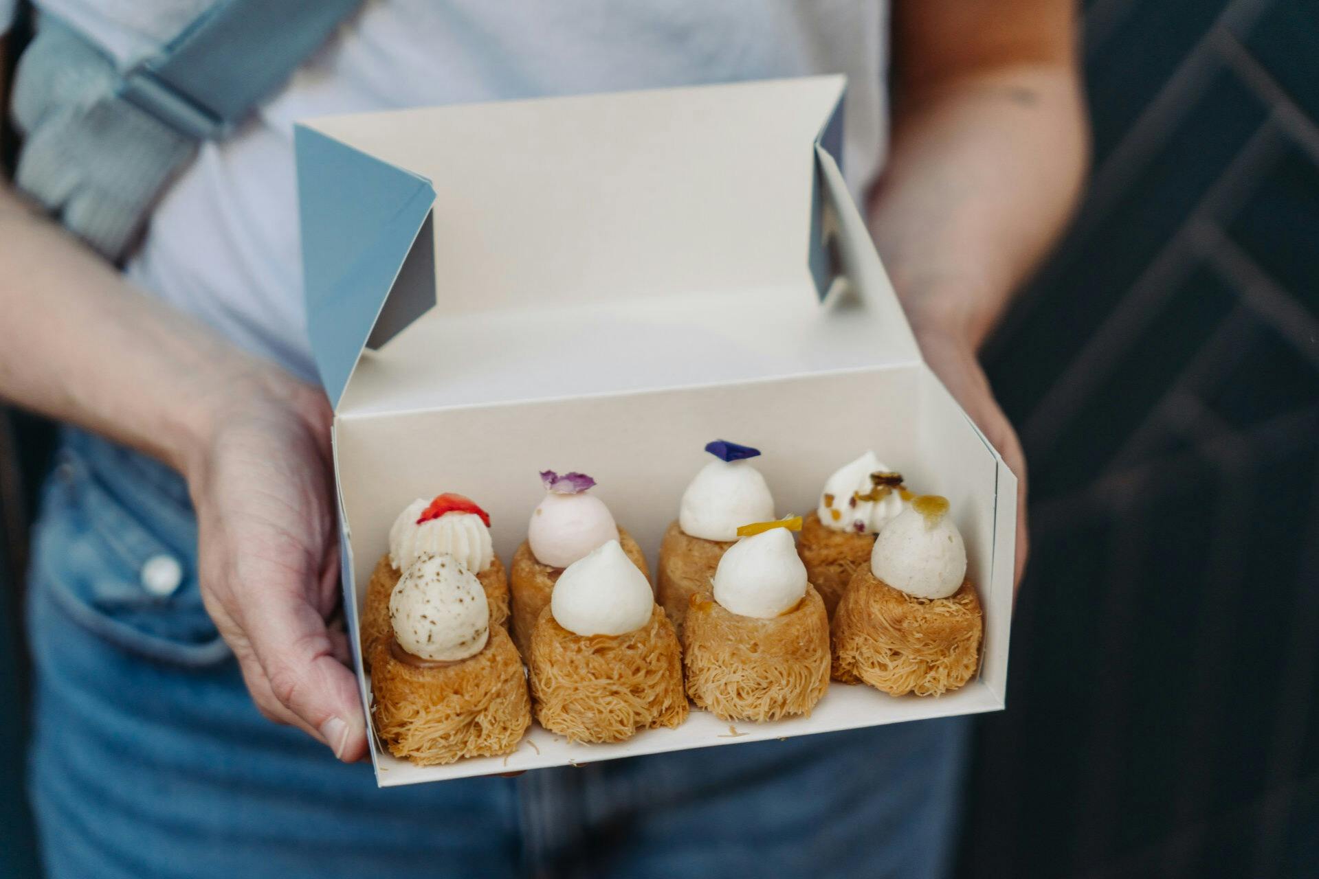 A person holding a box containing six pastries topped with cream and garnishes, each placed in a lined arrangement.