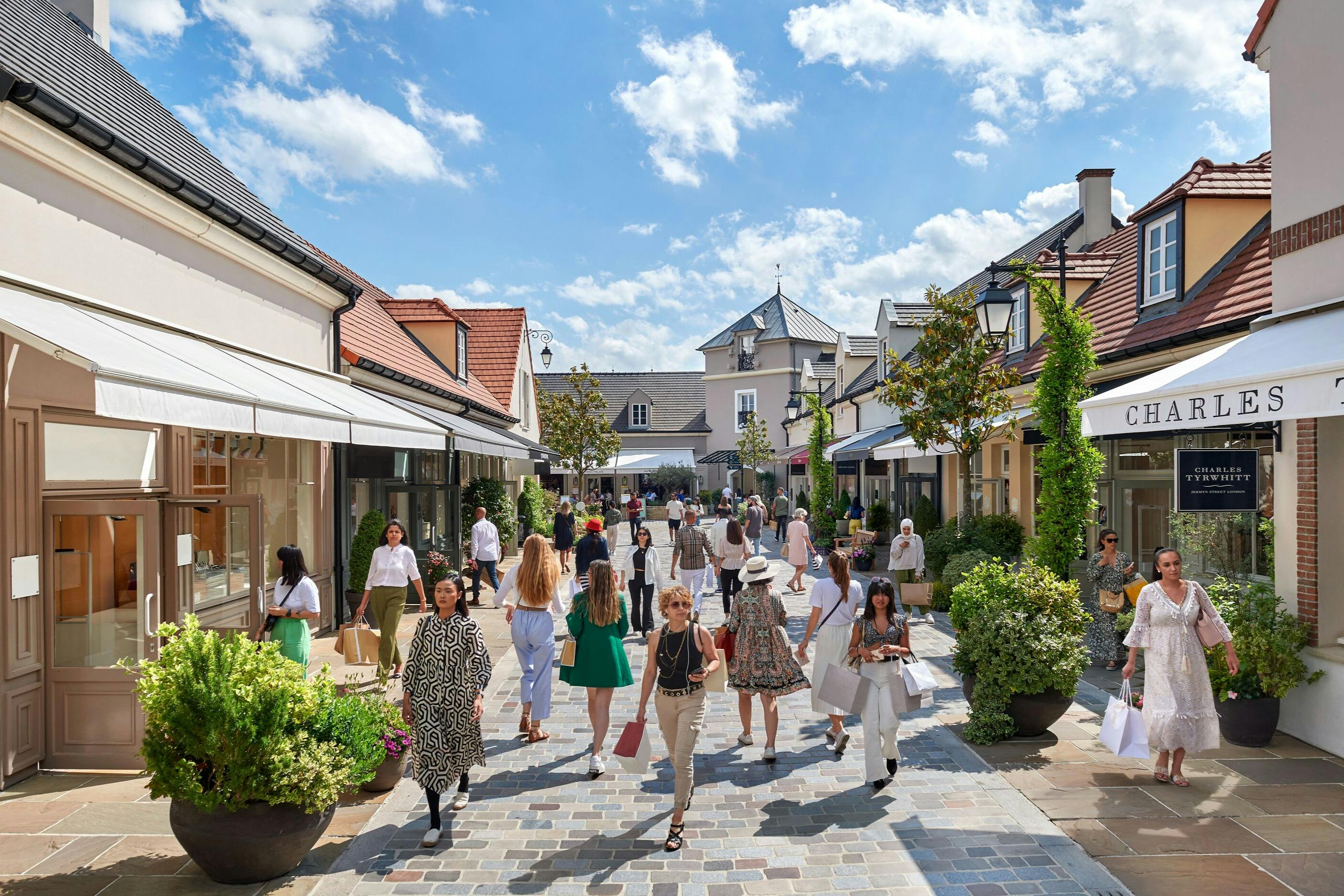 Ludzie spacerują i robią zakupy wzdłuż uroczej, otwartej ulicy handlowej z butikami, zielenią i niebieskim niebem z chmurami nad głową.