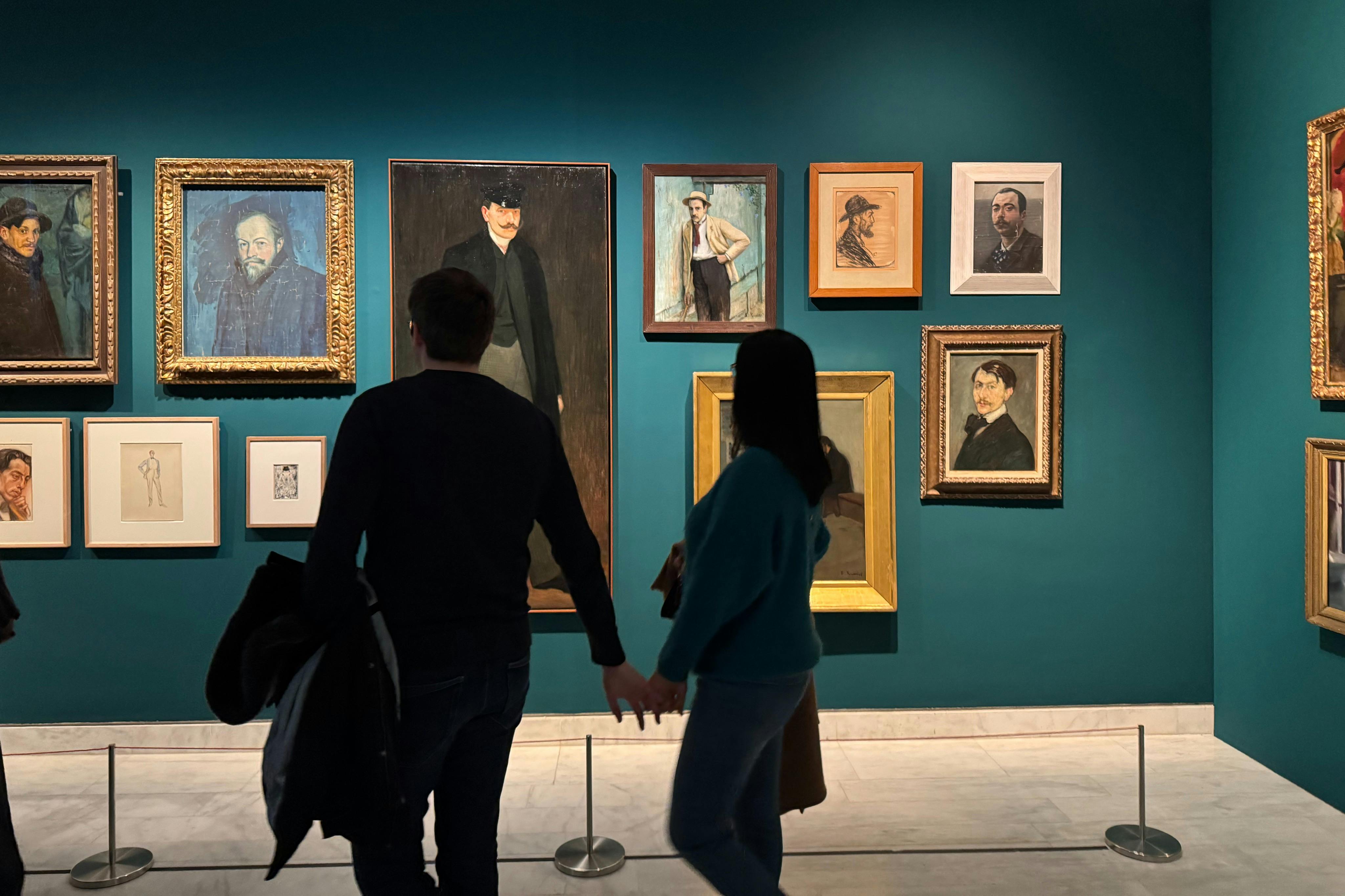 Une galerie faiblement éclairée aux murs d'un bleu sarcelle profond présente une collection des premiers portraits de Picasso.