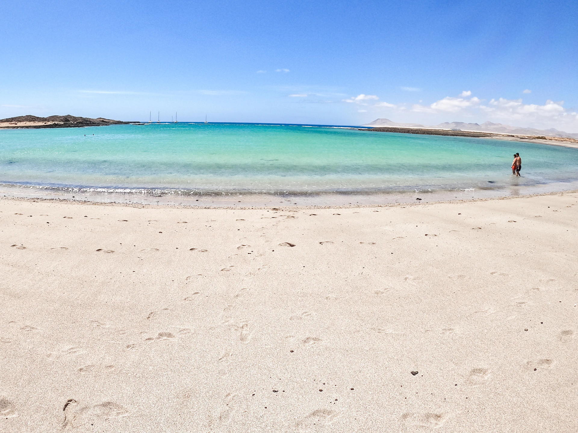 A serene beach with smooth sand, clear turquoise water, and a bright blue sky with light clouds.