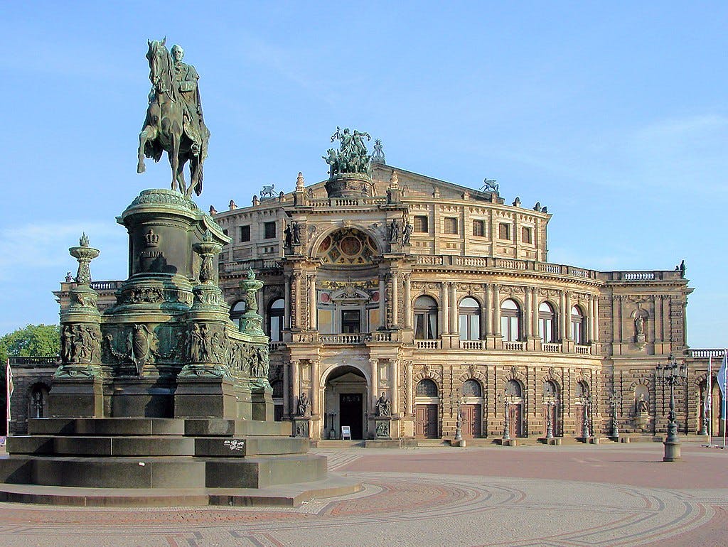 Equestrian statue in front of a detailed, historic building with arched windows and ornate sculptures, under a clear blue sky.