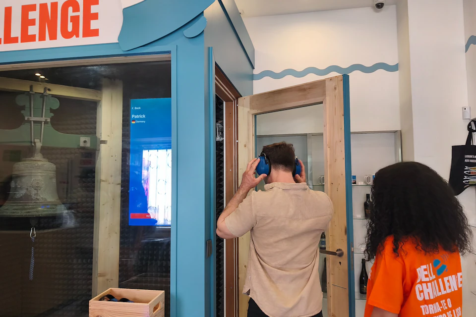 A person wearing blue headphones stands near a soundproof booth in a modern room, with another person nearby.