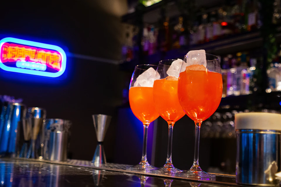 Three glasses with orange cocktails and ice cubes on a bar counter, with a neon sign and bottles in the background.