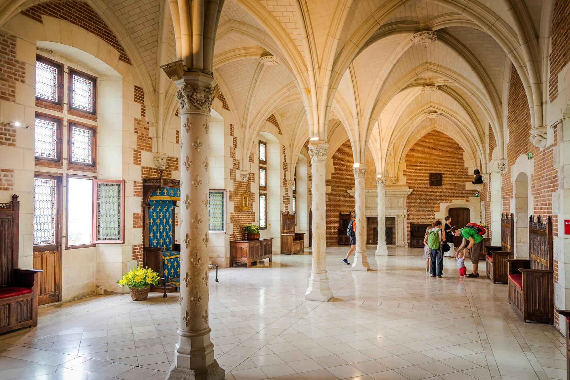 Una espaciosa sala medieval con techos abovedados, columnas y visitantes que contemplan el interior. El mobiliario incluye una chimenea y sillas.