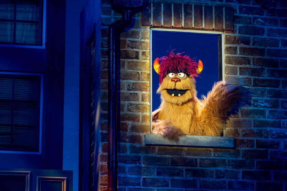 A puppet with orange fur, red hair, and yellow horns looks out from a brick-framed window in a dimly lit scene.