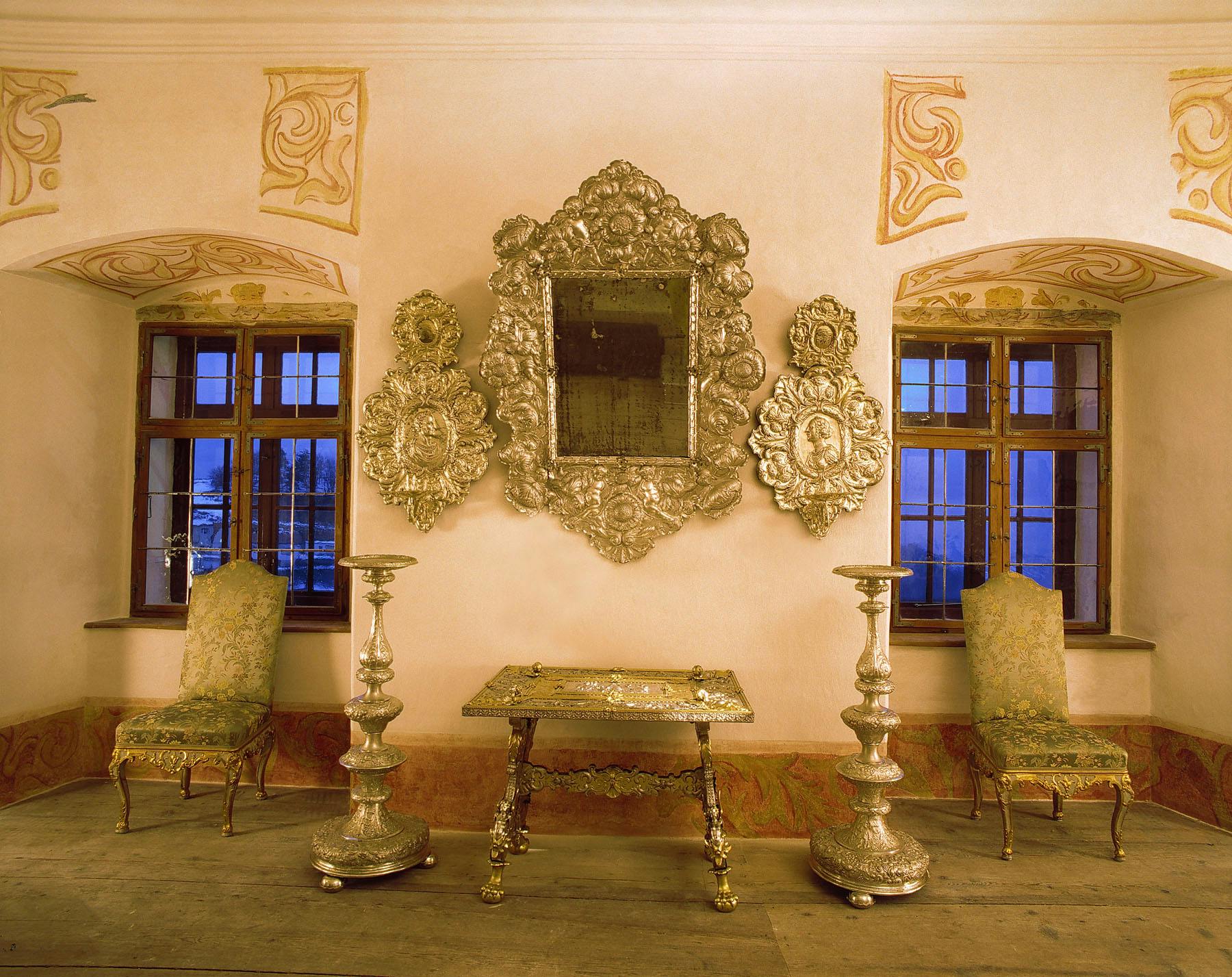 房间装饰华丽，有两扇高大的窗户、一面精雕细琢的镜子、配套的墙面装饰、金色扶手椅、一张小桌子和烛台。