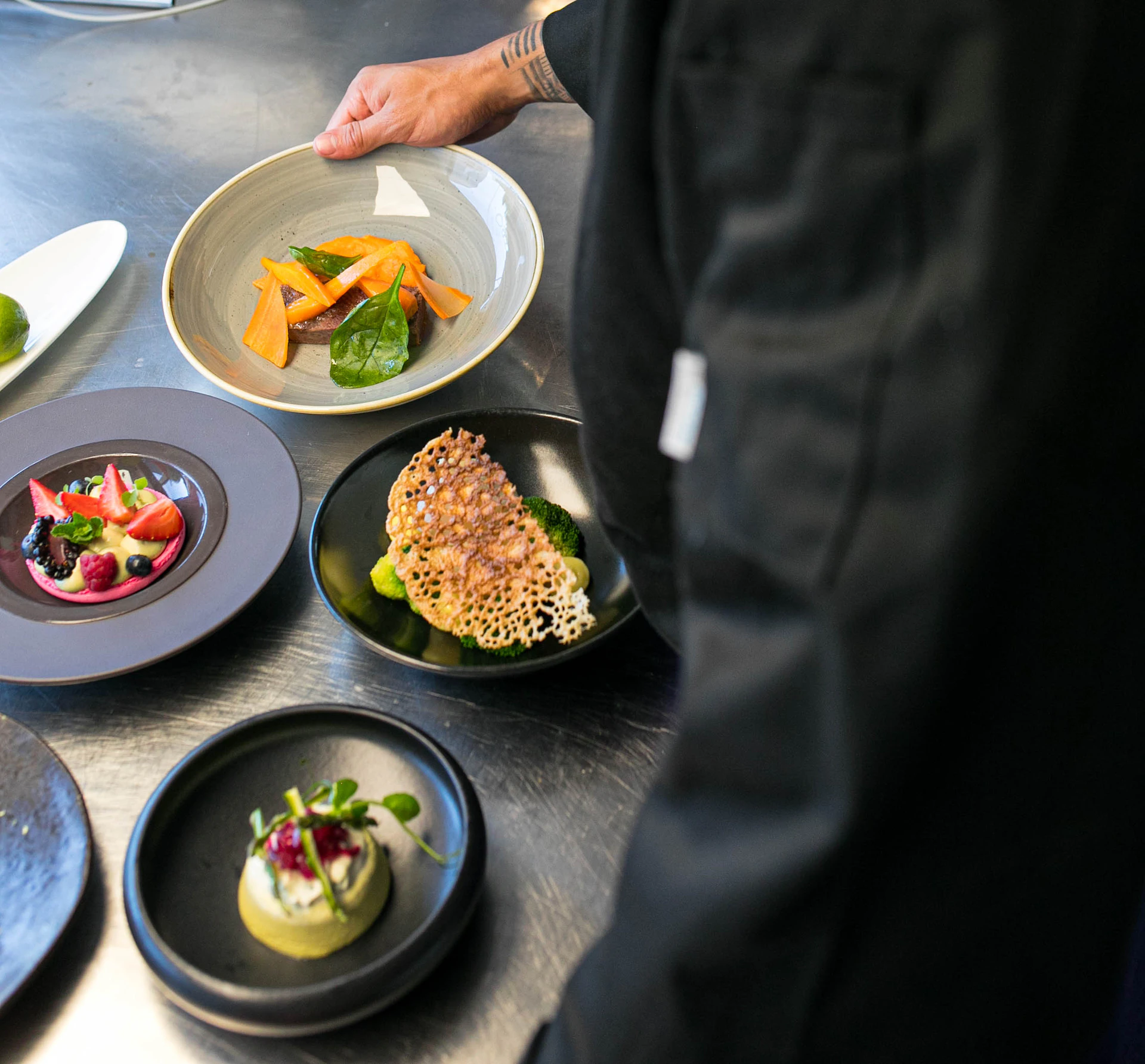 A chef in a black uniform arranges three gourmet dishes with vegetables, garnishes, and intricate plating on a metal surface.