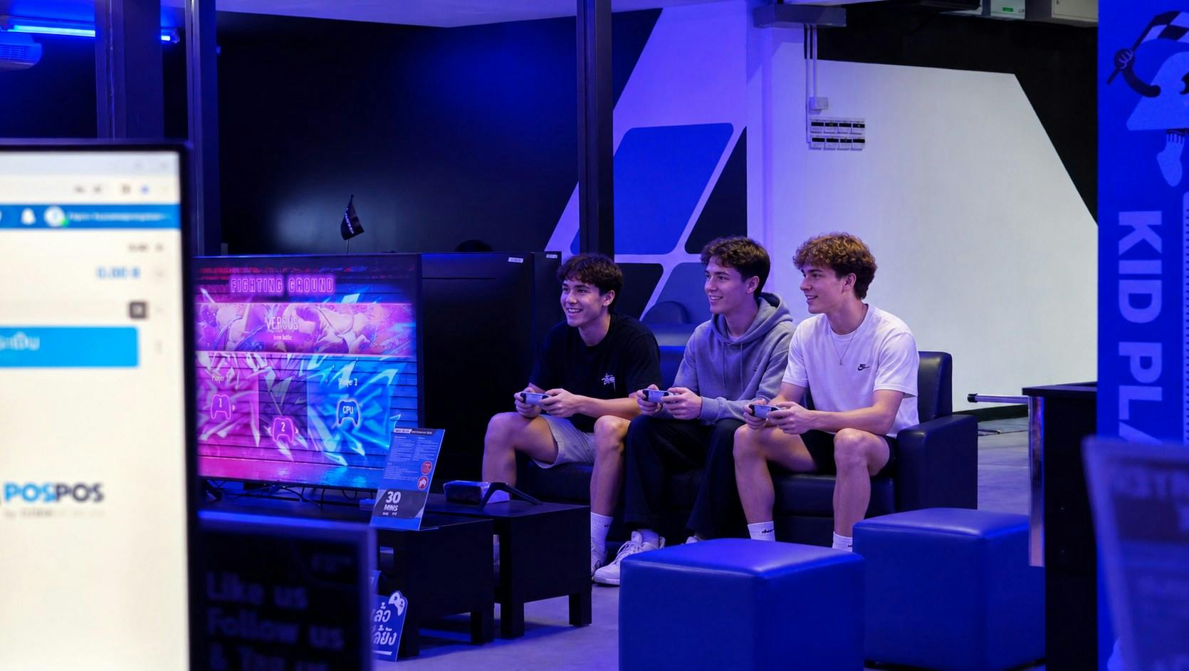 Three young men are sitting on a couch in a gaming lounge, holding controllers and smiling while playing video games.