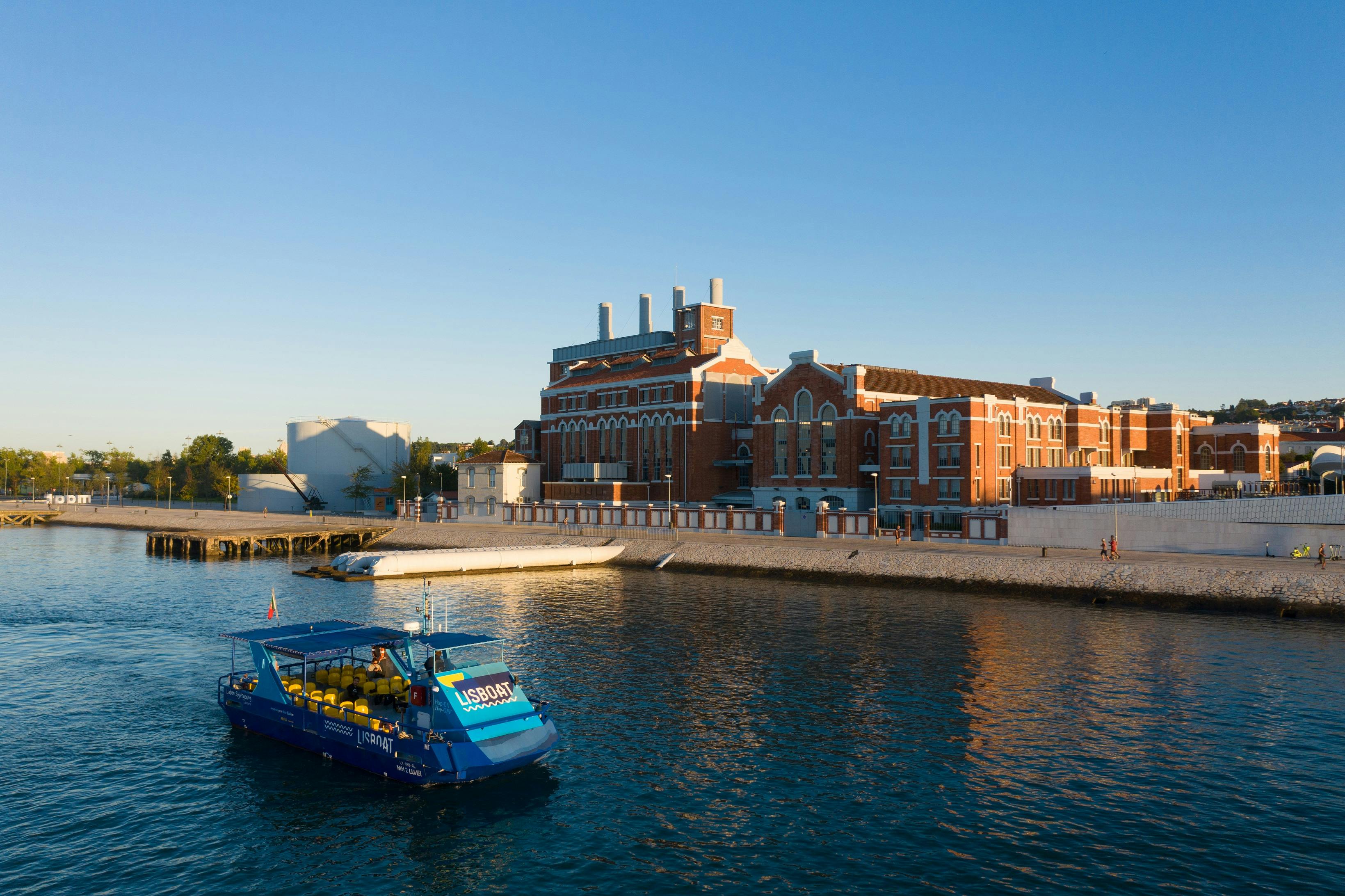 "USBOAT" 가 프린트된 파란색 투어 보트가 맑고 푸른 하늘 아래 물가의 크고 유서 깊은 붉은 벽돌 건물을 지나고 있습니다.